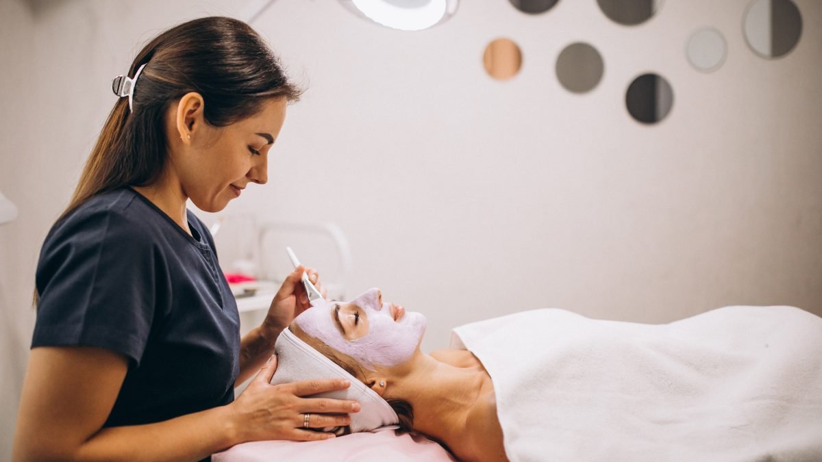 Cosmetóloga aplicando una solución exfoliante en el rostro de una clienta con los ojos cerrados